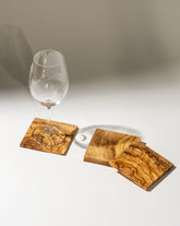 Three square olive wood coasters displayed on a white background, with a wine glass placed on top of one, highlighting their natural grain and functionality.